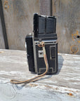 A vintage black and silver folding camera with a textured grip and a worn leather strap, photographed against a weathered wooden background.