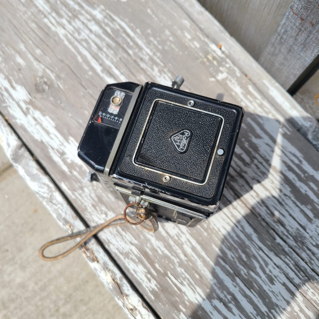 An antique black Rolle Rux camera with a textured leather front cover and silver metal accents, resting on a weathered, peeling white-painted wooden surface.