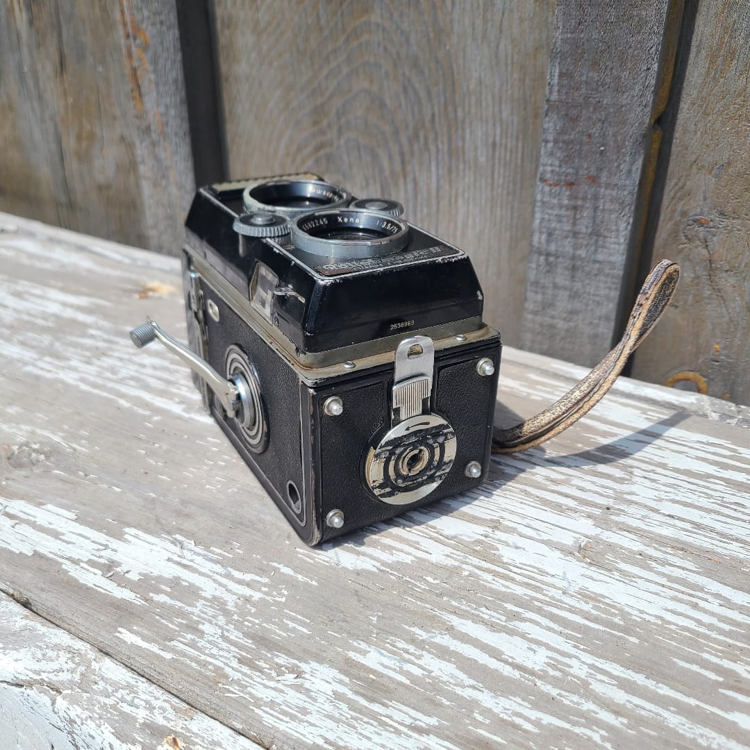 A vintage black and silver double-lens reflex camera with a leather strap, photographed at an angle against a weathered, peeling white-painted wooden surface.
