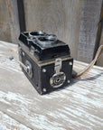 A vintage black and silver double-lens reflex camera with a leather strap, photographed at an angle against a weathered, peeling white-painted wooden surface.