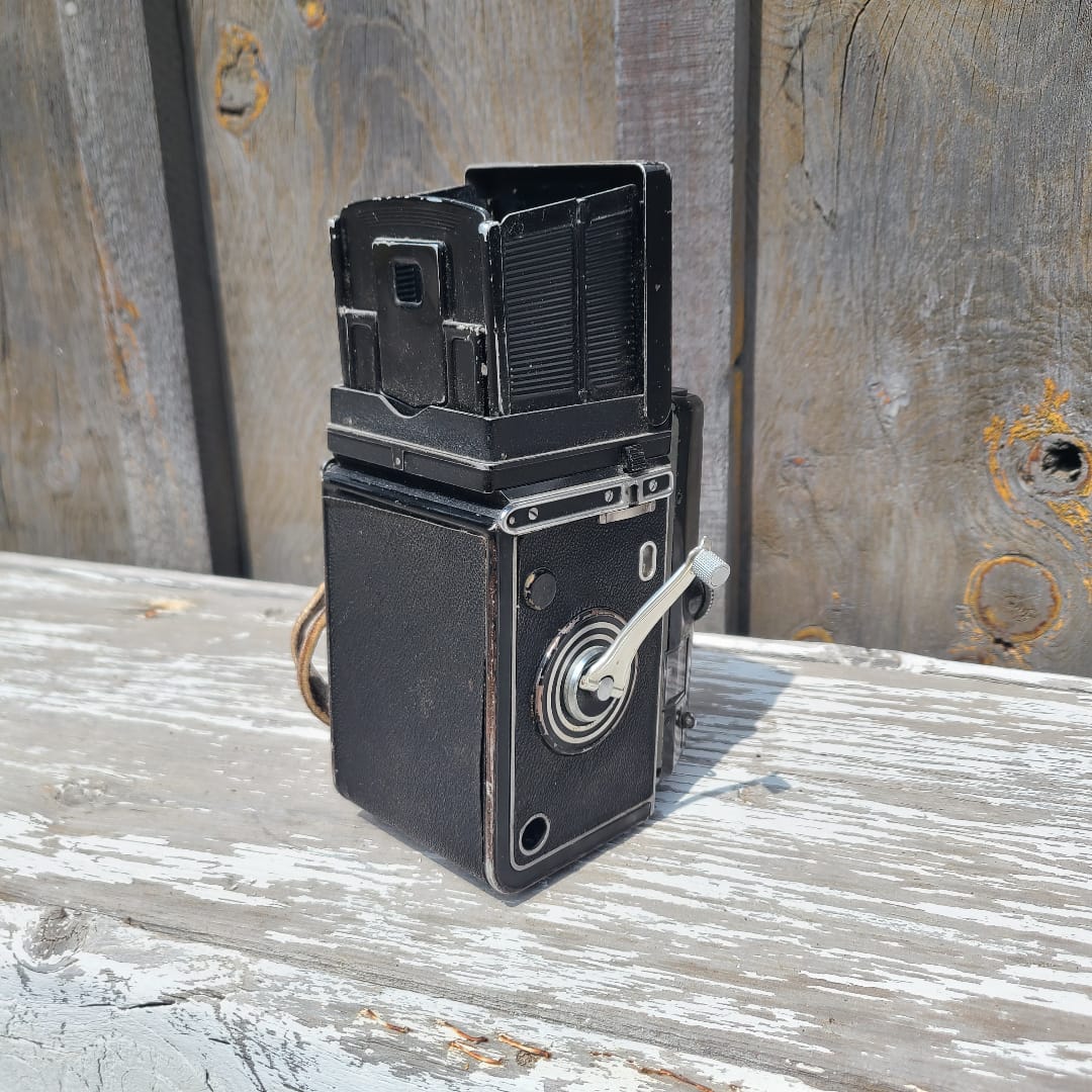 A vintage black metal camera body with a white hand crank handle, photographed mounted against weathered wooden boards.