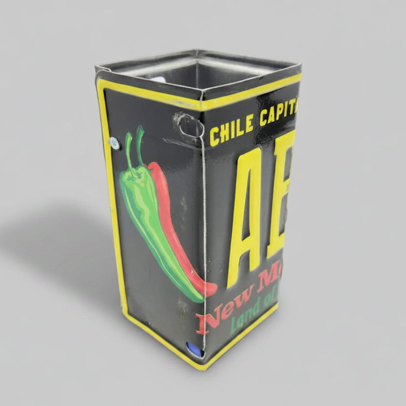 A repurposed New Mexico license plate pencil holder with black, yellow, and green colors, featuring text and a chili pepper graphic.